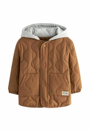 Dziecko ubrane w brązową pikowaną kurtkę z kapturem, czarne spodnie cargo i białe trampki, stojące w pomieszczeniu na jasnych schodach.