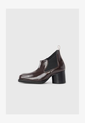 Bottine en cuir verni marron avec des panneaux élastiques sur les côtés, bout arrondi et talon épais noir. Présente une languette à l'arrière.