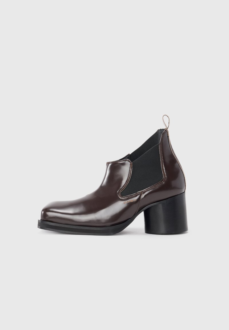 Bottine en cuir verni marron avec des panneaux élastiques sur les côtés, bout arrondi et talon épais noir. Présente une languette à l'arrière.