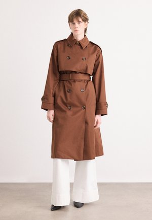 Bruine trenchcoat van textuurstof, met double-breasted knopen, bijpassende riem en een brede kraag. Model draagt witte wijdere broeken.