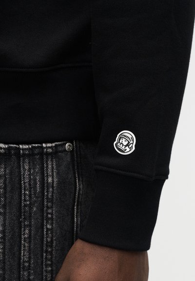 Μαύρο φούτερ με ριμπ μανσέτες, με μικρό λευκό λογότυπο αστροναύτη στο αριστερό μανίκι. Φορεμένο με σκούρο, ψηφιακό τζιν με σχέδια.