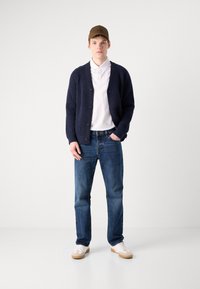 Μπλε πλεκτή ζακέτα, λευκό πουκάμισο με γιακά, μπλε τζιν και λευκά αθλητικά παπούτσια με καφέ σόλες. Το μοντέλο φοράει ένα καφέ καπέλο με λογότυπο.