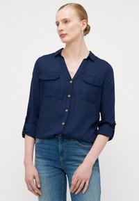 Chemise bleu marine à boutons avec manches retroussables, deux poches poitrine et boutons dorés. Fabriquée dans un tissu léger au toucher lisse.