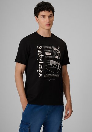 Młody mężczyzna z kręconymi ciemnymi włosami, ubrany w czarny t-shirt z nadrukiem "Sunday League" i niebieskie spodnie cargo, stoi na tle jednolitego szarego tła.