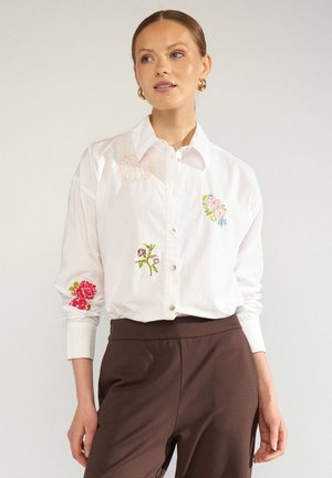 Femme portant une chemise blanche boutonnée avec une broderie florale et un pantalon marron taille haute, debout devant un fond uni.