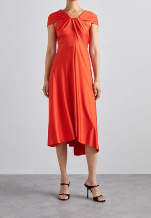 Vrouw draagt een knielange, feloranje jurk met off-shoulder mouwen en asymmetrische zoom, gecombineerd met zwarte sandalen met hoge hakken.