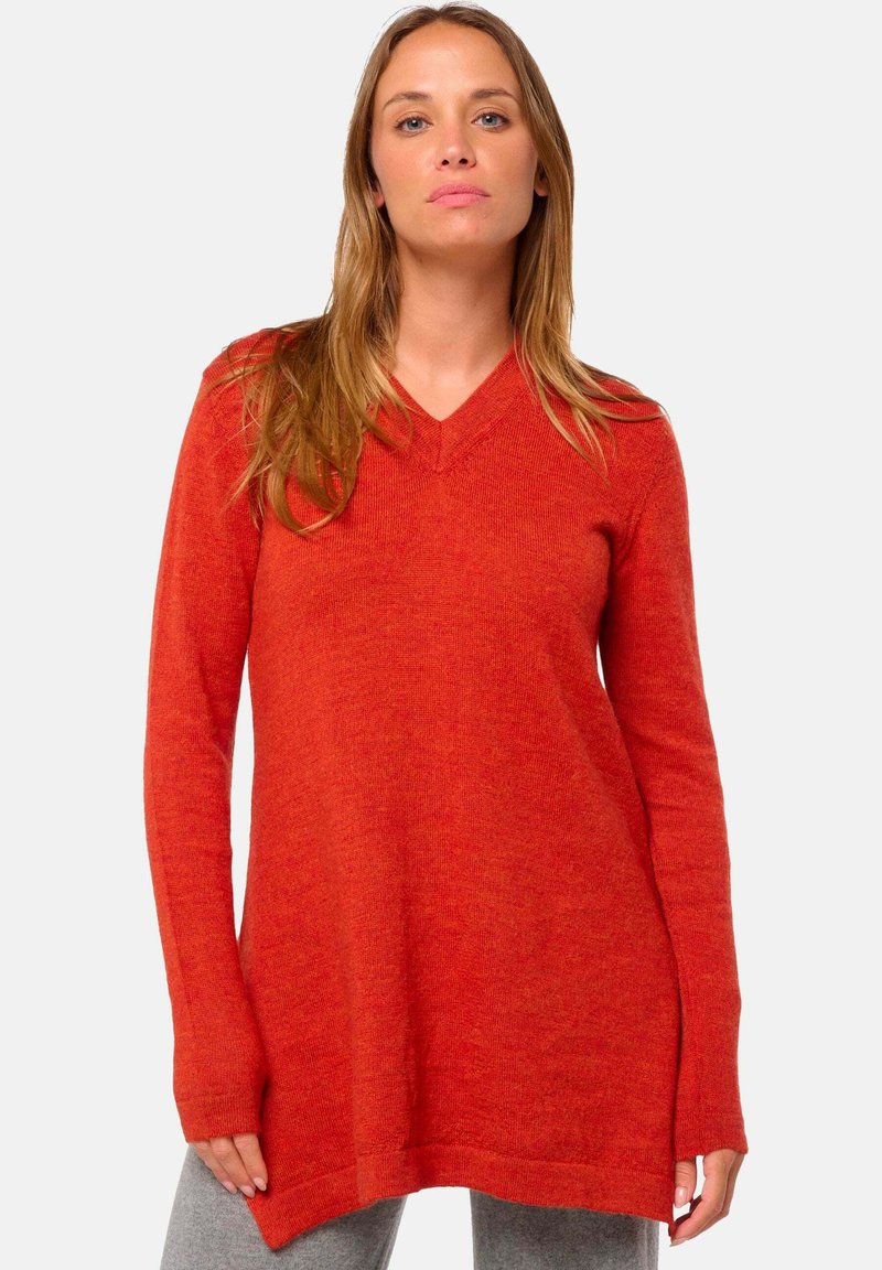 Femme portant un pull rouge à manches longues et décolleté en V, se tenant contre un fond gris clair uni.