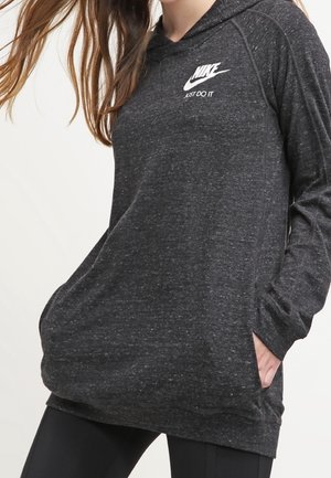 Modelka nosząca ciemnoszarą bluzę z kapturem Nike z logo "Just Do It", ręce w przedniej kieszeni, w połączeniu z czarnymi legginsami.