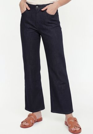 Kobieta ubrana w ciemne, szerokie dżinsy z wysokim stanem, z rękami w kieszeniach i brązowe sandały na odkryte palce, stojąca na białym tle.