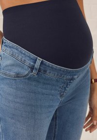 Jeans de maternitate cu un brâu elastic de culoare albastru închis și denim de culoare albastru deschis. Vedere în prim-plan care subliniază brâul și buzunarele.