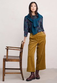 Camisa de manga larga con estampado floral combinada con pantalones de pana anchos en color mostaza y botas de cordones marrón rojizo. Bufanda de punto azul caída sobre los hombros.