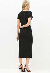 Schwarzes Midi-Kleid mit kurzen Ärmeln, taillierter Silhouette und glatter Textur. Kombiniert mit schwarzen High-Heel-Sandalen. Dezente seitliche Nähte sichtbar.