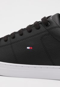 Sneaker in pelle nera con finitura testurizzata, suola bianca e lacci marroni. Presenta un piccolo emblema rosso, bianco e blu sul lato.