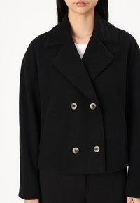 Black double-breasted jacket with a collar, made of a textured fabric, featuring two light-colored buttons and wide sleeves.
