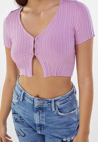 Top corto de canalé rosa con escote en V, mangas cortas y botones frontales, combinado con jeans azules desgastados que presentan diseños de texto mezclados.