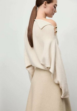 Suéter beige de gran tamaño con textura de canalé, cuello ancho y botones en los hombros. Combinado con una falda beige a juego, de tejido suave y diseño minimalista.
