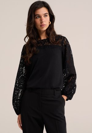 Femme aux longs cheveux foncés portant un chemisier en dentelle noire avec des manches transparentes et un pantalon noir, se tenant devant un fond neutre.