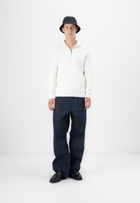 Sudadera blanca con cremallera, jeans de pierna ancha azul oscuro, sombrero tipo cubo negro y zapatos negros. Diseño simple con colores y texturas contrastantes.