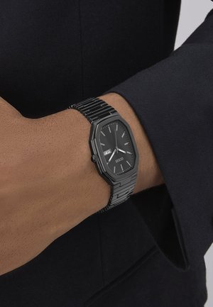 Black octagonal watch on a wrist, featuring a black dial, silver hour markers, and a metal link bracelet. Date display at 3 o'clock.