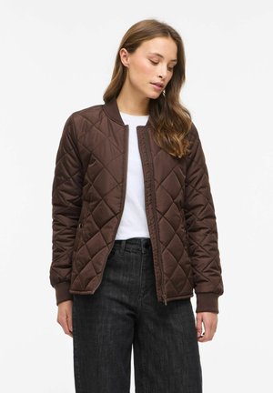 Femme aux longs cheveux bruns portant une veste matelassée marron par-dessus une chemise blanche et un jean noir, regardant vers le bas les yeux fermés.