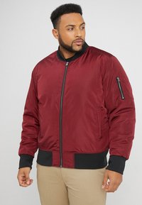 Giubbotto bomber bordeaux con zip frontale, polsini neri e vita. Presenta tasche laterali e una tasca con zip sulla manica. Tessuto liscio e resistente.