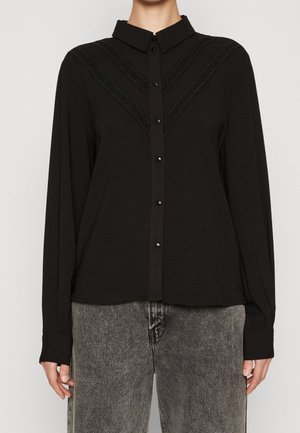 Zwarte blouse met lange mouwen en knopen aan de voorkant, versierd met een kanten V-patroon over de borst, gecombineerd met vervaagde grijze jeans.