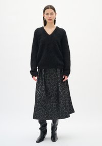 Frau in einem flauschigen schwarzen V-Ausschnitt-Pullover, einem grauen und schwarzen Leo-Print-Midirock, schwarzen spitz zulaufenden Stiefeln und silbernen Creolen.