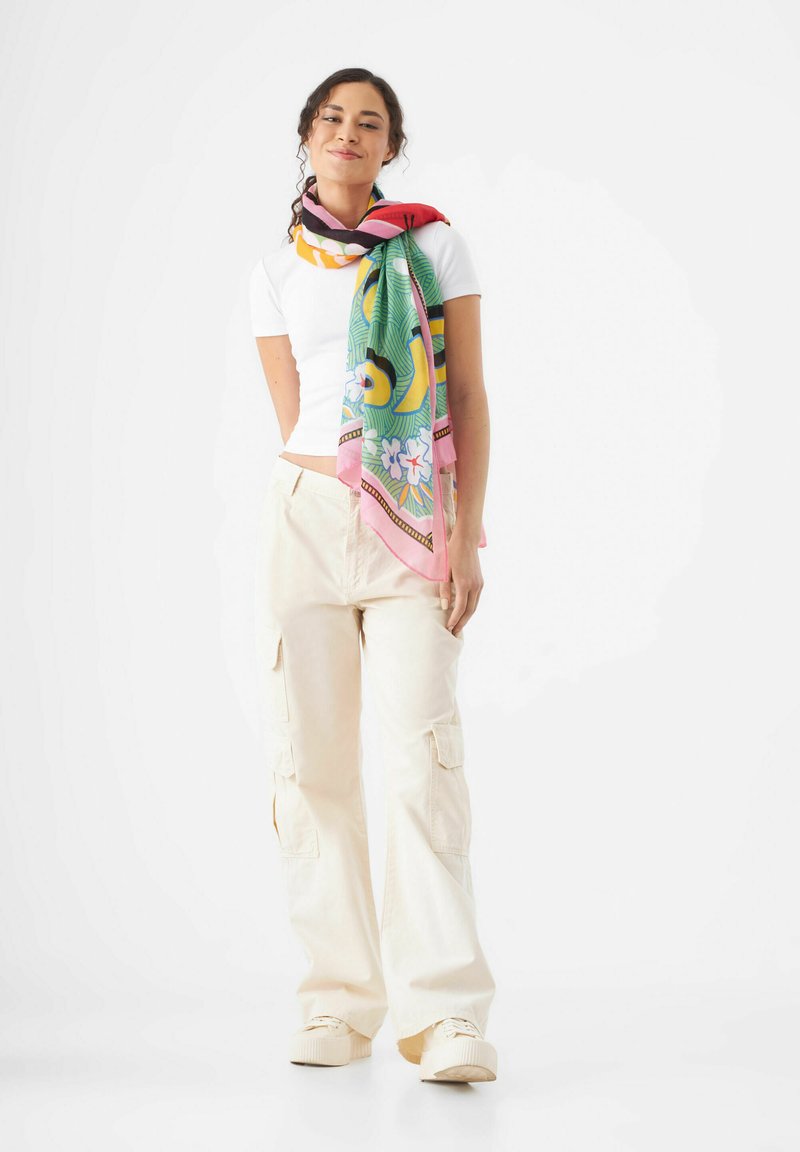 Foulard en soie coloré avec des motifs floraux et abstraits, enroulé autour du cou, associé à un haut blanc ajusté et un pantalon cargo crème.