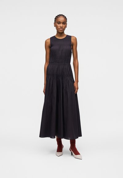 Femme debout portant une robe midi noire sans manches avec des détails froncés, associée à des chaussettes couleur rouille et des chaussures blanches à talons.