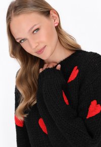 Schwarzer gestrickter Pullover mit großen roten Herzmustern, mit einem gerippten Ausschnitt und Bündchen. Model trägt rosa gestreifte Ohrringe.