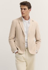 Beige strukturierter Blazer mit zwei Vordertaschen, einem einzigen Knopfverschluss und einem kleinen Pin-Detail am Revers, getragen über einem weißen Hemd.