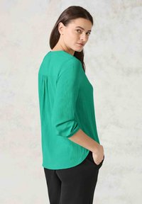 Grön texturerad blus med långa ärmar, avslappnad passform och rundad nederkant. Har plisseringar bak och uppvikta ärmslut.