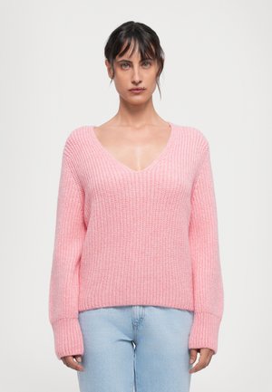 Mulher com cabelo escuro vestindo um suéter rosa de malha grossa em V e jeans claros, de pé contra um fundo branco simples.