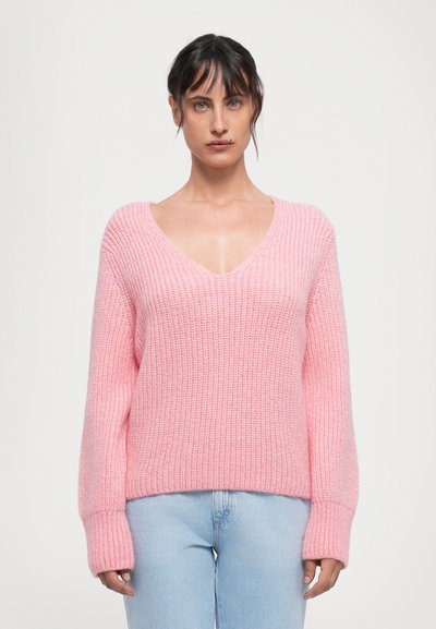 Mujer con cabello oscuro vistiendo un suéter de punto grueso rosa con cuello en V y jeans azul claro, de pie contra un fondo blanco liso.