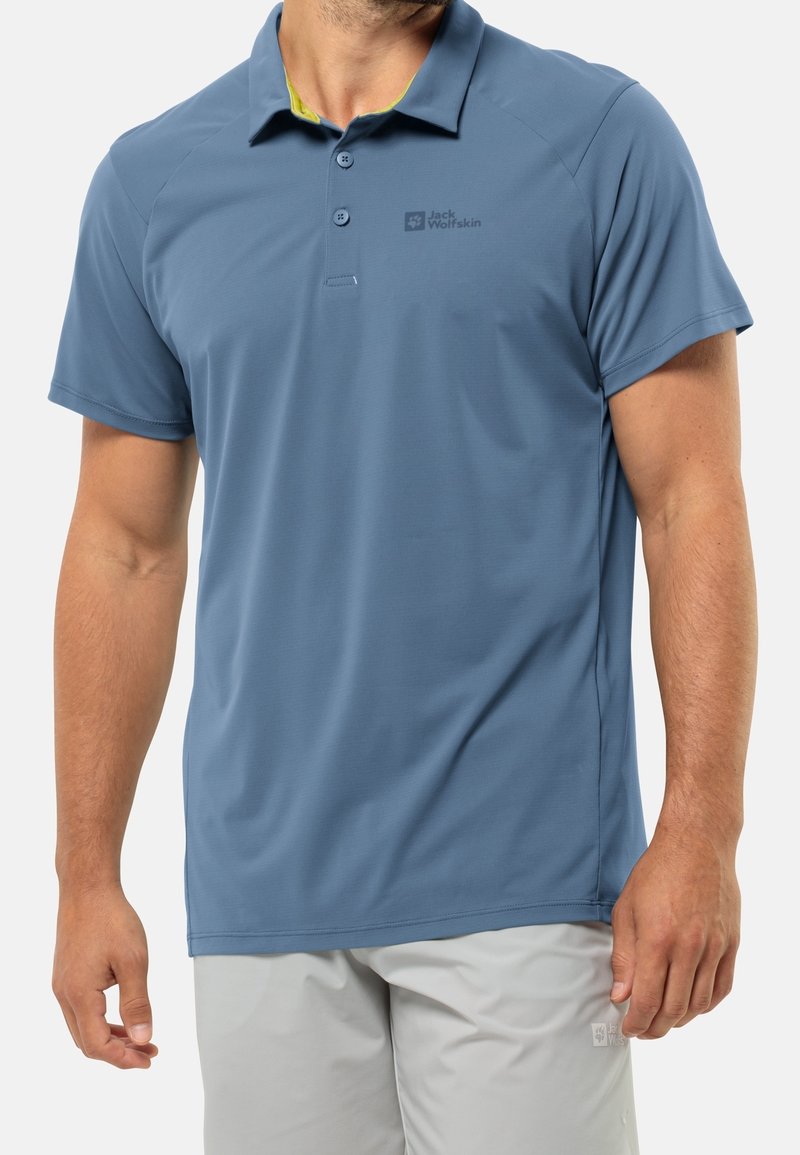 Homme portant un polo bleu à manches courtes et un short gris clair, debout avec les bras détendus le long du corps.