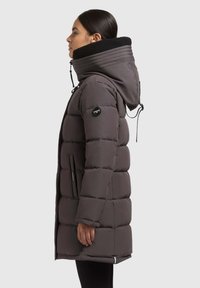 Abrigo acolchado gris con cuello alto, cordón elástico y bolsillos laterales. Presenta un forro interior negro texturizado y una marca en la manga.