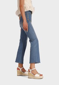 Jeans in denim con taglio a zampa, di colore azzurro chiaro, con dettagli in bottone dorato. Abbinati a sandali con zeppa bianchi con un dettaglio a fibbia rotonda.