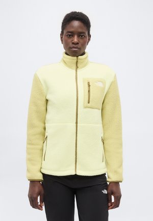 Személy előre nézve, világos sárga polár dzsekit visel, melyen cipzáros mell- és oldalzsebek találhatók, valamint fekete nadrágot.