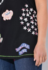 Camiseta negra con gráficos de estrellas coloridas, el texto "Sé feliz" y "Amor divertido y paz", con costuras verdes a lo largo del dobladillo.