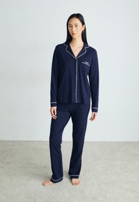 Conjunto de pijama azul-marinho com mangas longas e calças. Apresenta um acabamento em branco ao longo da gola, punhos e bainha. Inclui um bolso no peito.