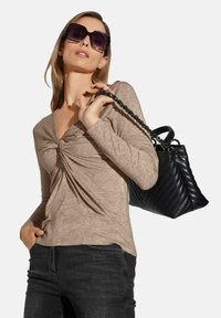 Hellbraunes Langarmoberteil mit einem verdrehten Vorderteil-Design, kombiniert mit dunklen Jeans und einer schwarzen, gesteppten Handtasche mit Kettenriemen. Große schwarze Sonnenbrille.