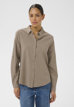 Femme aux cheveux foncés portant une chemise beige à manches longues boutonnée et un jean bleu, debout devant un fond clair uni.