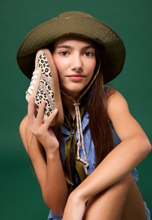Jeune femme portant un chapeau vert tient une basket à motif léopard près de son visage sur un fond vert uni.