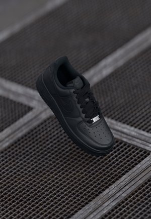 Čierne tenisky Nike Air Force 1 plávajúce nad kovovou mriežkou so perforovaným dizajnom a štítkom na šnúrkach s nápisom "AF1."