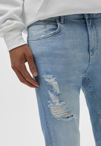 Světle modré opotřebované džíny se třepícími se roztrženími na levém stehně, doplněné bílou mikinou s dlouhým rukávem. Hladká denní textura.