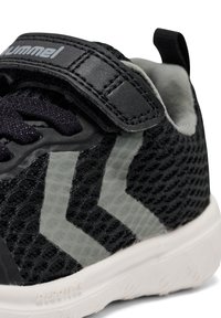 Schwarze Sneaker mit Mesh-Obermaterial, grauen Chevron-Akzenten, gepolstertem Kragen und verstellbarem Riemen. Leichte Gummisohle mit strukturiertem Grip.