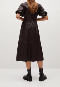 Vestido de cuero sintético marrón con mangas cortas abullonadas y cintura definida, que cuenta con una falda hasta la rodilla y se combina con botas negras hasta el tobillo.