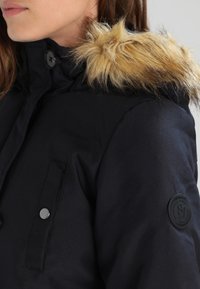 Kvinna som bär en svart vinterjacka med en knappad ficka, en knappad hög krage och en tanfärgad huva med fuskpäls.