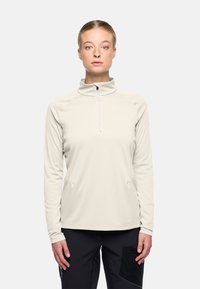 Vrouw in een lichtbeige langharige rits top en zwarte broek, rechtopstaand en recht vooruit kijkend met een neutrale uitdrukking.