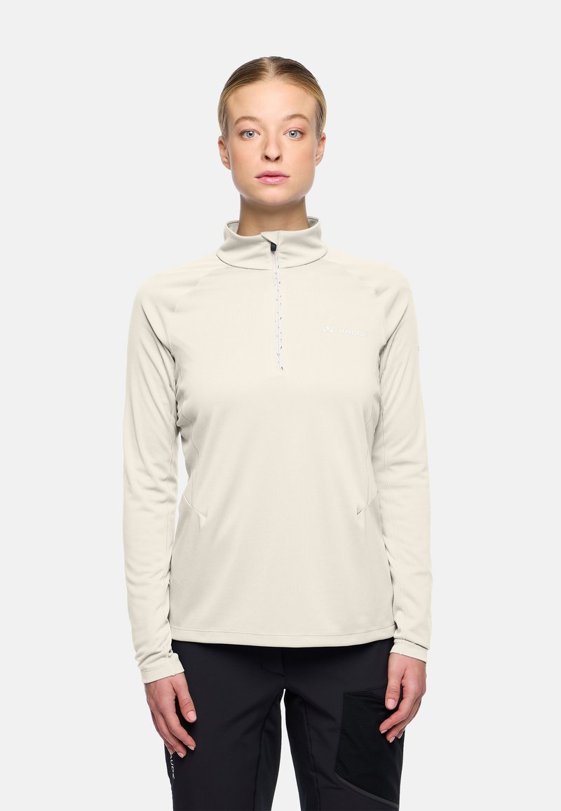 Vrouw in een lichtbeige langharige rits top en zwarte broek, rechtopstaand en recht vooruit kijkend met een neutrale uitdrukking.
